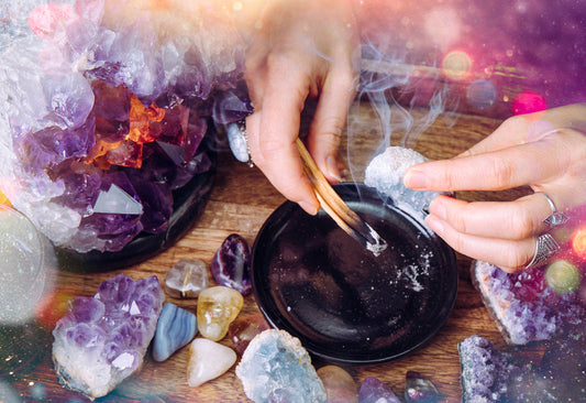 Hands holding a burning incense stick over a black bowl surrounded by amethyst crystals and other colorful stones on a wooden surface.