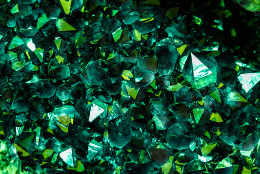 Close-up view of multiple dark green crystals with sharp, reflective facets and geometric shapes, creating a dense cluster with a shimmering surface